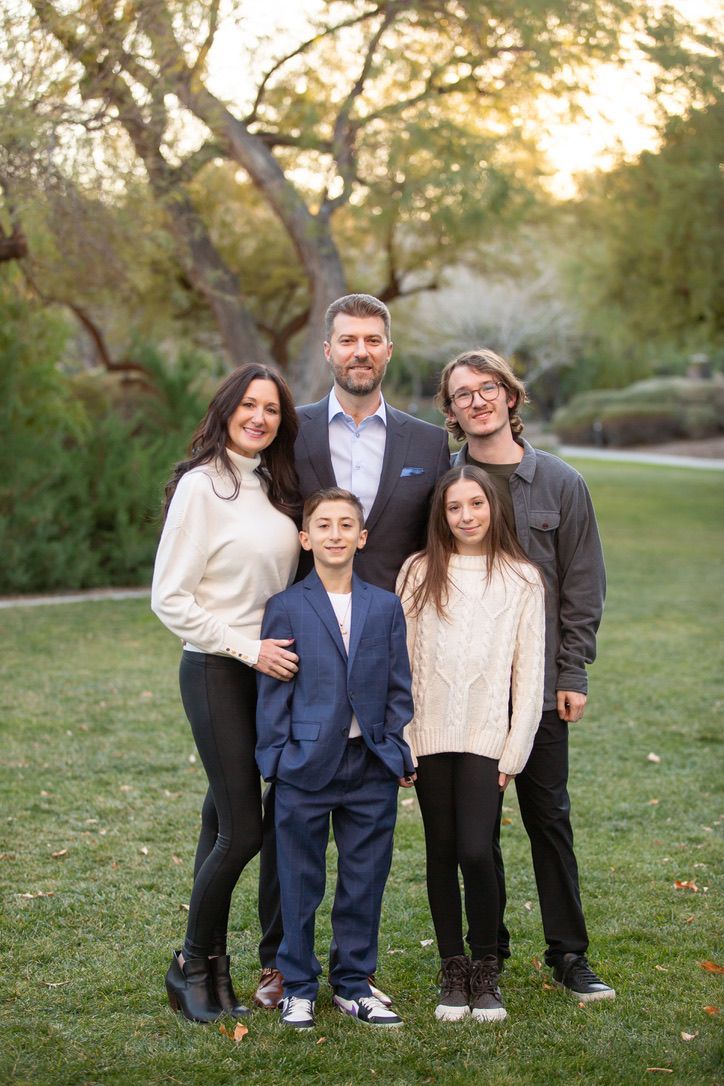 A family poses for a portrait in a grassy park at sunset, featuring a mix of casual and formal attire.