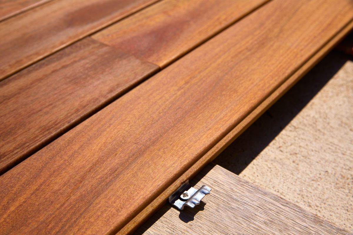 High-resolution image focusing on a detail of a wooden deck installation in Lubbock, TX. Rich, warm-toned wooden planks are laid out neatly side by side with the grains of the wood creating a natural pattern. A silver metal fastener, designed for deck assembly, connects two planks ensuring a seamless and secure fit. The fastener's sharp lines and industrial appearance contrast with the organic textures of the wood. The sun casts shadows accentuating the wood's texture and the fastener's silhouette.