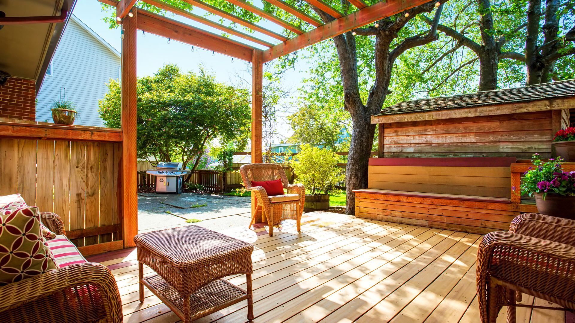 A charming wooden deck in Lubbock, TX, is furnished with wicker chairs and a matching coffee table. The area is covered by a pergola adorned with string lights, creating a cozy outdoor living space. Cushions with vibrant patterns add a welcoming touch.  Lush greenery surrounds the area, while a wooden bench, potted plants, and a BBQ grill enhance the functional and aesthetic aspects. The scene is framed by trees and a wooden fence, creating a serene and inviting environment for relaxation and outdoor gatherings.