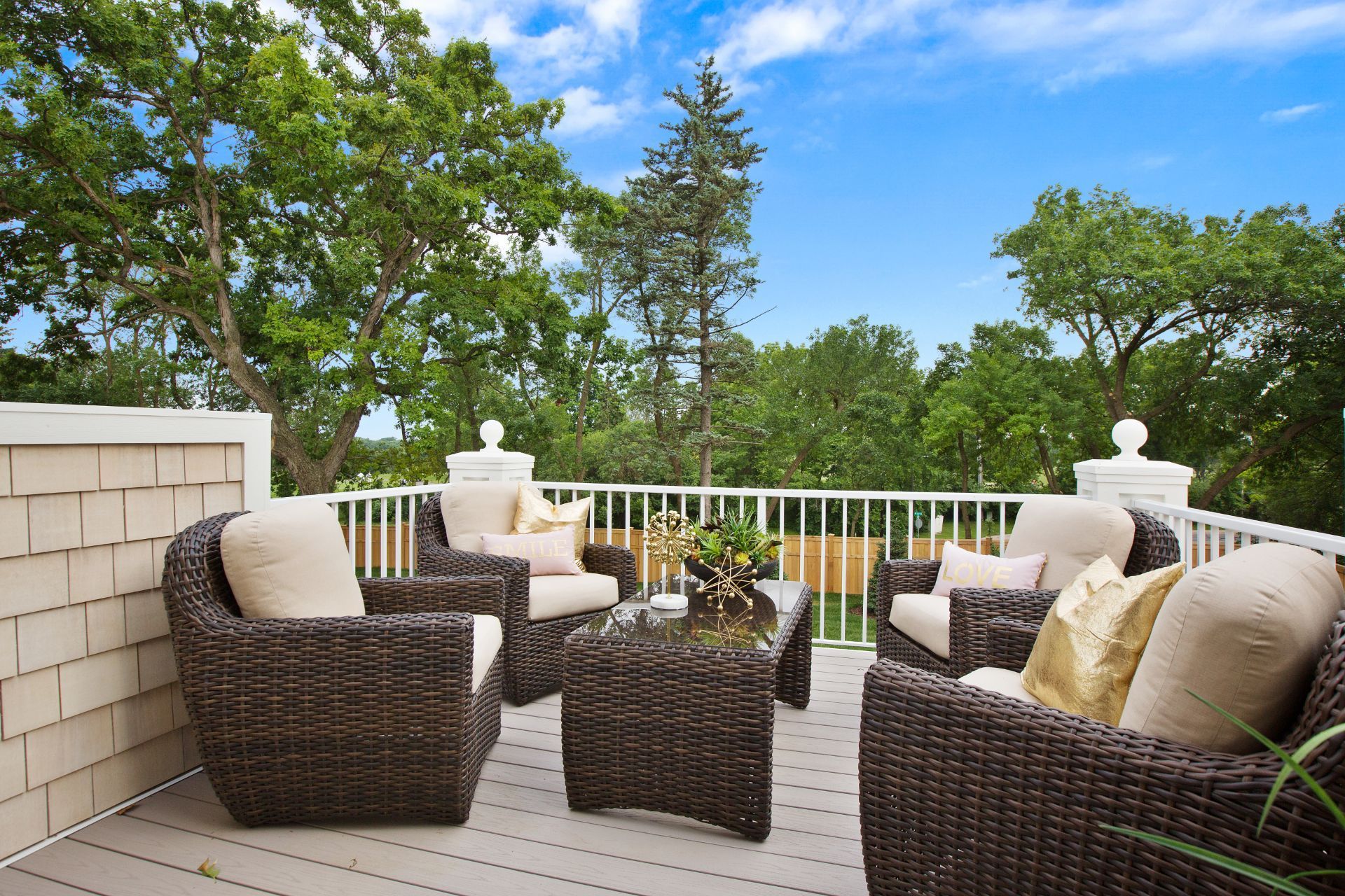 An inviting outdoor deck in Lubbock, TX, with a contemporary design featuring comfortable wicker furniture with plush cushions and decorative pillows. The setting includes a coffee table with a plant centerpiece, surrounded by mature trees under a clear blue sky. The white railing adds a classic touch to the deck, offering a cozy space for relaxation and enjoying the outdoors.