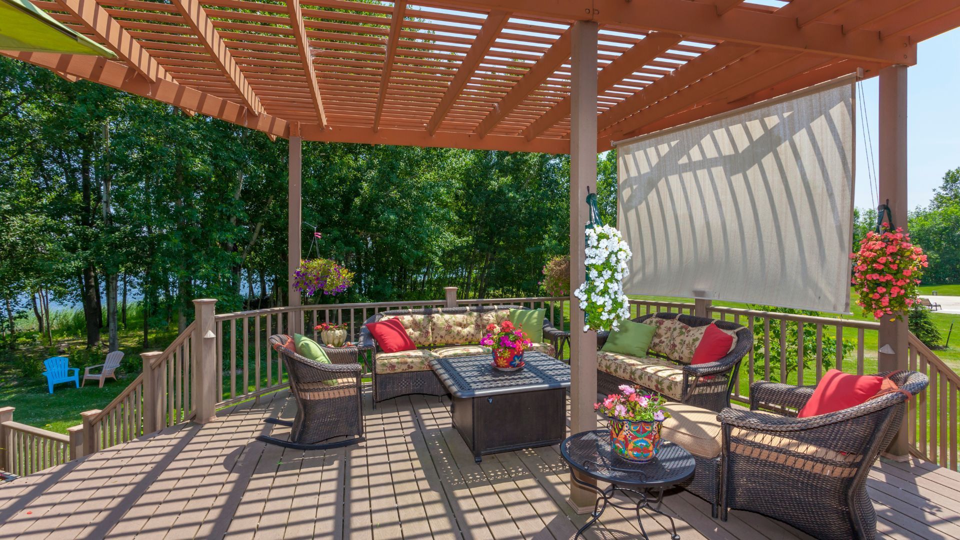 This inviting outdoor deck area in Lubbock, TX, features a luxurious pergola with lattice roofing, offering filtered sunlight over a cozy seating arrangement. The deck includes rattan furniture with patterned cushions and vibrant floral arrangements, enhancing the space's aesthetic. A retractable screen provides shade on bright days. The deck overlooks a lush backyard with mature trees and a glimpse of a calm lake, creating a tranquil retreat perfect for relaxation and entertainment.