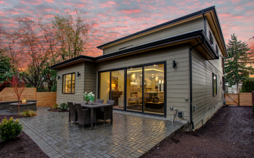 A photo of a beautiful backyard patio built by Hub City Deck Pros. The patio has a firepit built to the left and a nice table in the center. The sun is setting in the photo, so the sky looks blue and red.