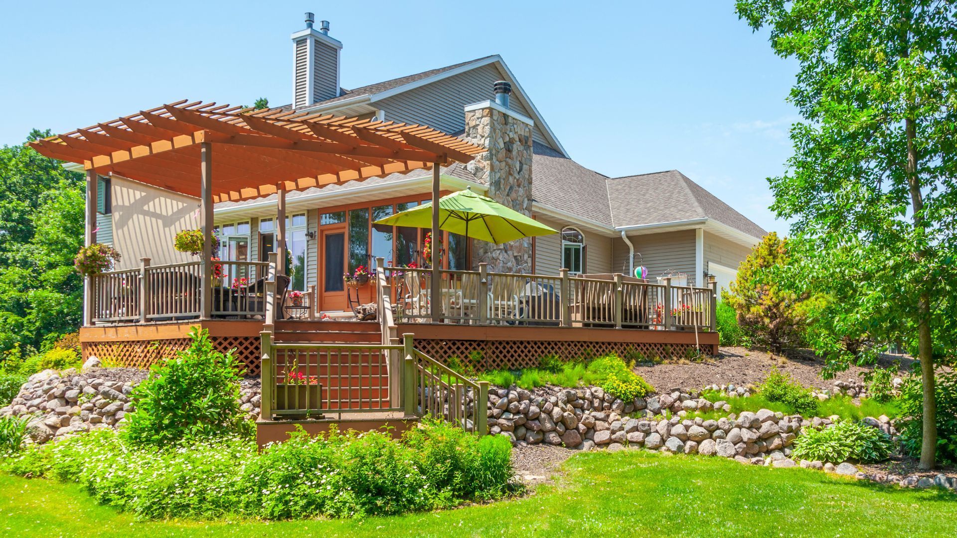 This image showcases a beautifully crafted deck and pergola in Lubbock, TX, adjacent to a stately home with a stone and siding exterior. The deck is equipped with a wooden pergola providing partial shade, while various potted plants and flowers enhance its cozy, inviting ambiance. The structure overlooks a well-maintained garden and lush greenery, offering a picturesque outdoor living space ideal for relaxation and entertainment.