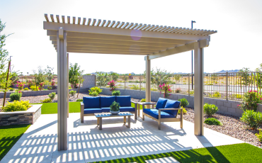 A stunning photo of a pergola in Lubbock, TX. The Pergola is made completely out of wood. The pergola has patio furniture sitting underneath the structure. The patio is surrounded by bright green grass and shrubbery.