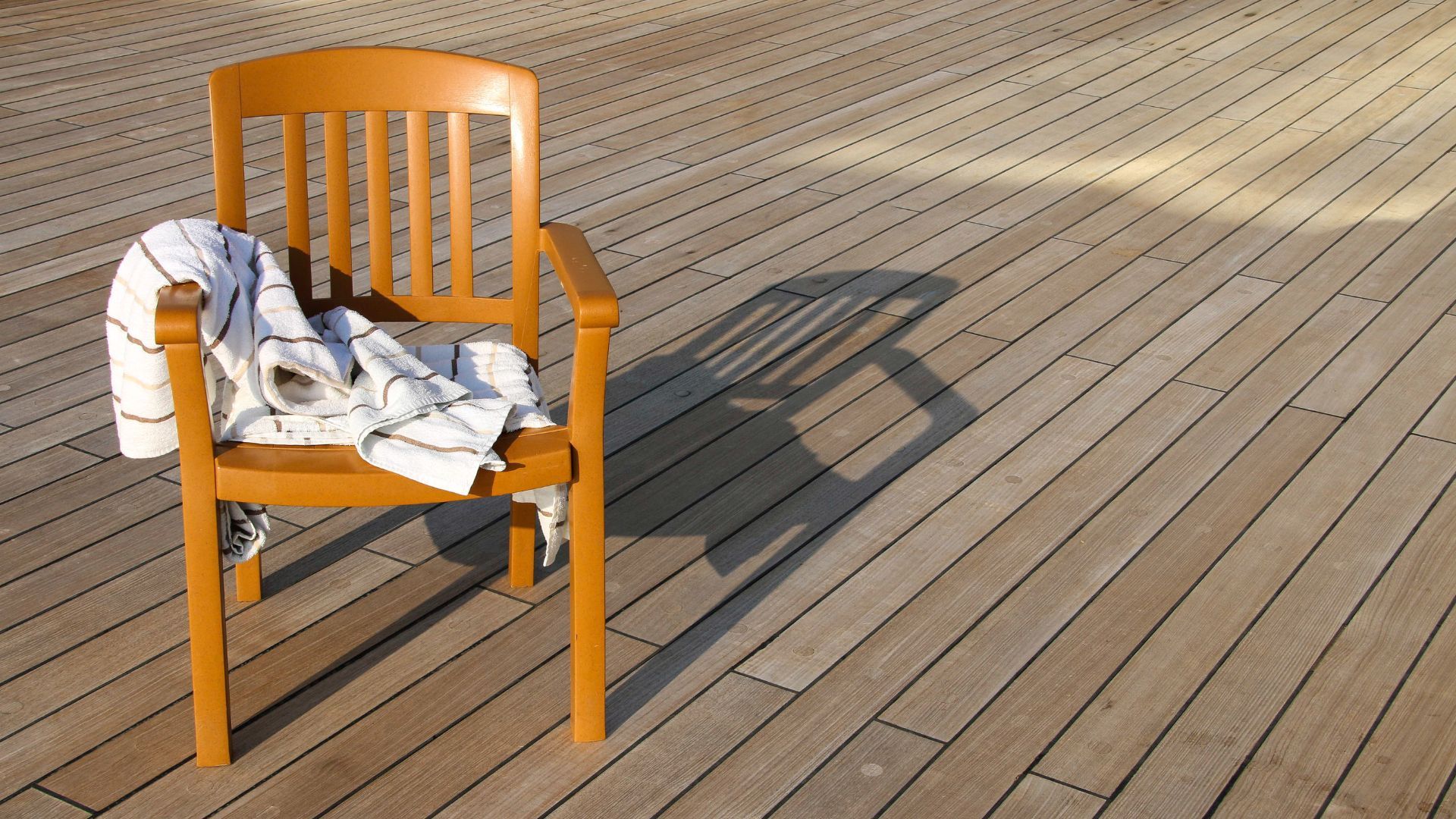 A simple, serene scene on a wooden deck in Lubbock, TX, featuring a single wooden chair with a striped cloth draped over it. The deck's planks are arranged in a neat, parallel fashion, creating a clean, minimalist look. The chair casts a long shadow, suggesting a sunny day. The composition highlights a sense of quiet relaxation, perfect for enjoying a peaceful moment outdoors. This setup reflects the laid-back, comfortable ambiance typical of a Lubbock home.