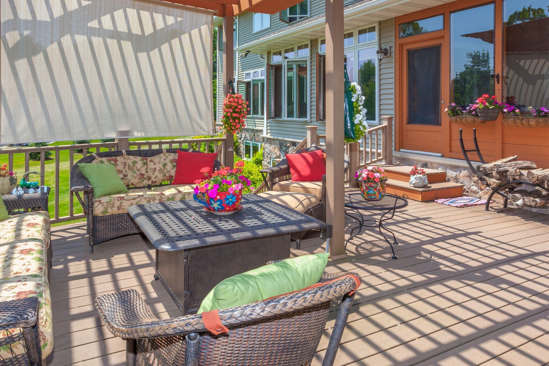 A welcoming deck in Lubbock, TX, features a comfortable wicker seating arrangement with patterned cushions and colorful throw pillows. Blooming potted plants add a dash of color against the backdrop of the home's exterior. The space is shaded by a retractable awning, creating a relaxing environment for enjoying the outdoors, complete with a fire pit table and a rocking chair, inviting leisure and entertainment.