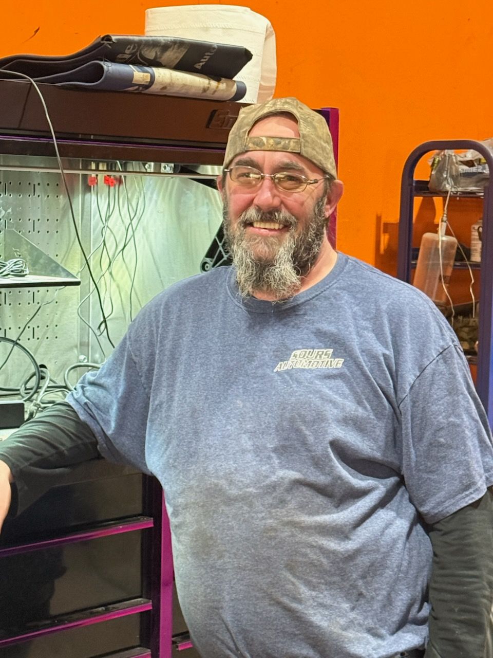 Man with beard and glasses wearing a cap and blue shirt, smiling in an orange-walled shop.