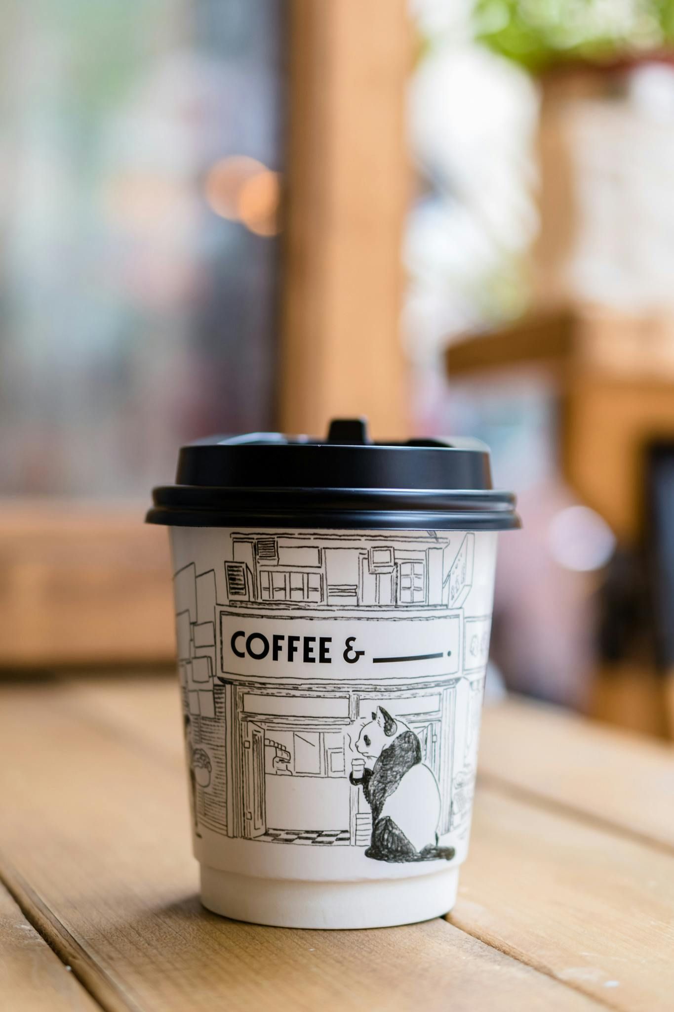 Coffee cup with black lid on a wooden table, drawing of coffee shop on the cup.