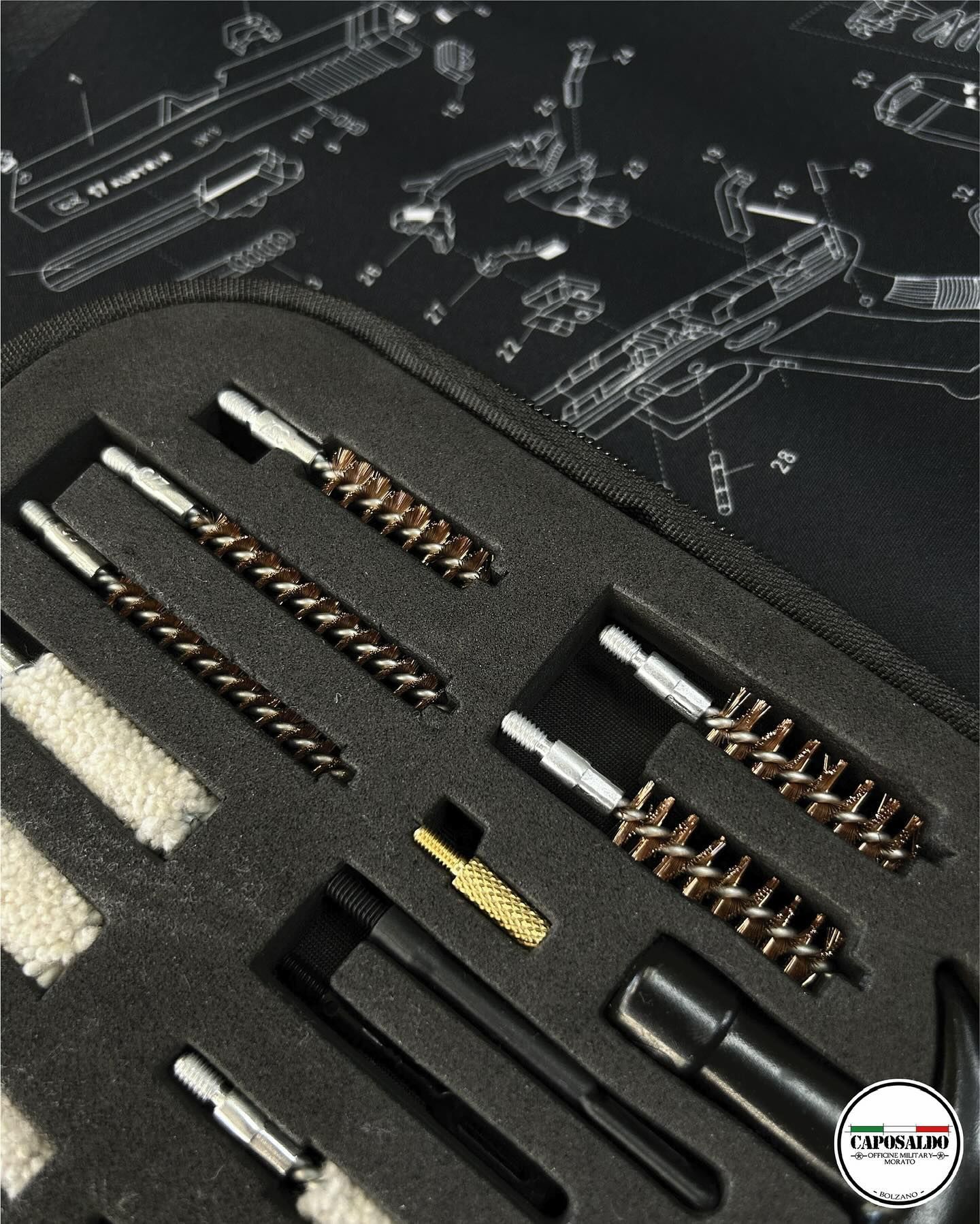 Custodia in schiuma nera con assortimento di spazzole, aste e piccoli attrezzi per la pulizia delle armi, su una superficie scura con motivo decorativo.