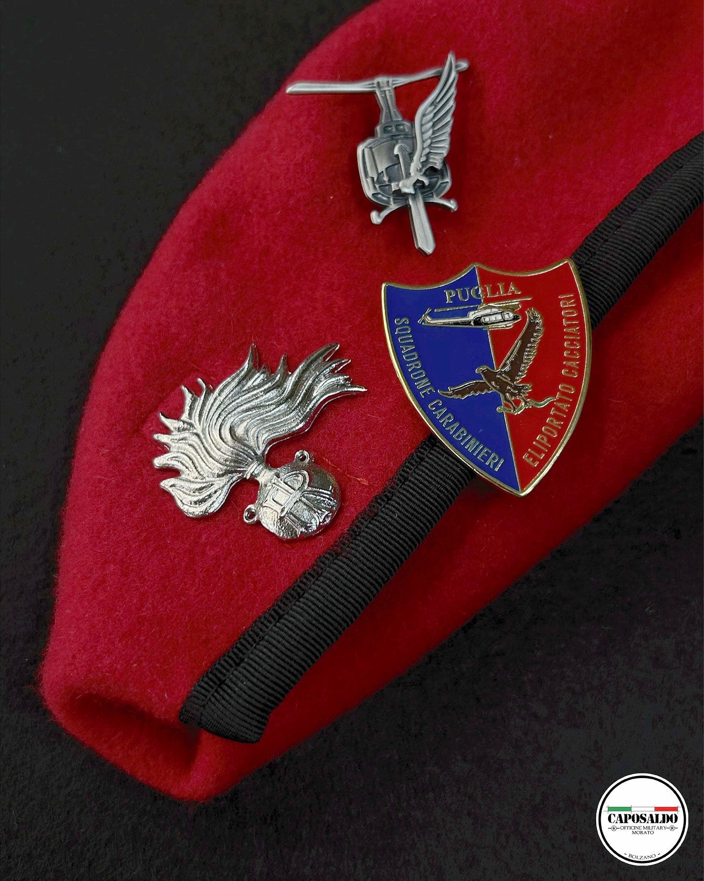 Berretto rosso con spille in stile militare e stemma a scudo blu su sfondo scuro.
