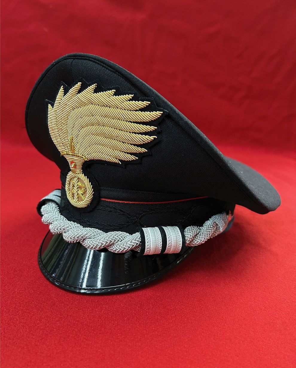 Berretto nero con visiera, emblema alato dorato e treccia argentata su sfondo di tessuto rosso.