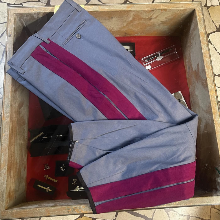 Pantaloni grigi piegati con pannelli laterali magenta in una cassa di legno, con piccole parti metalliche sotto