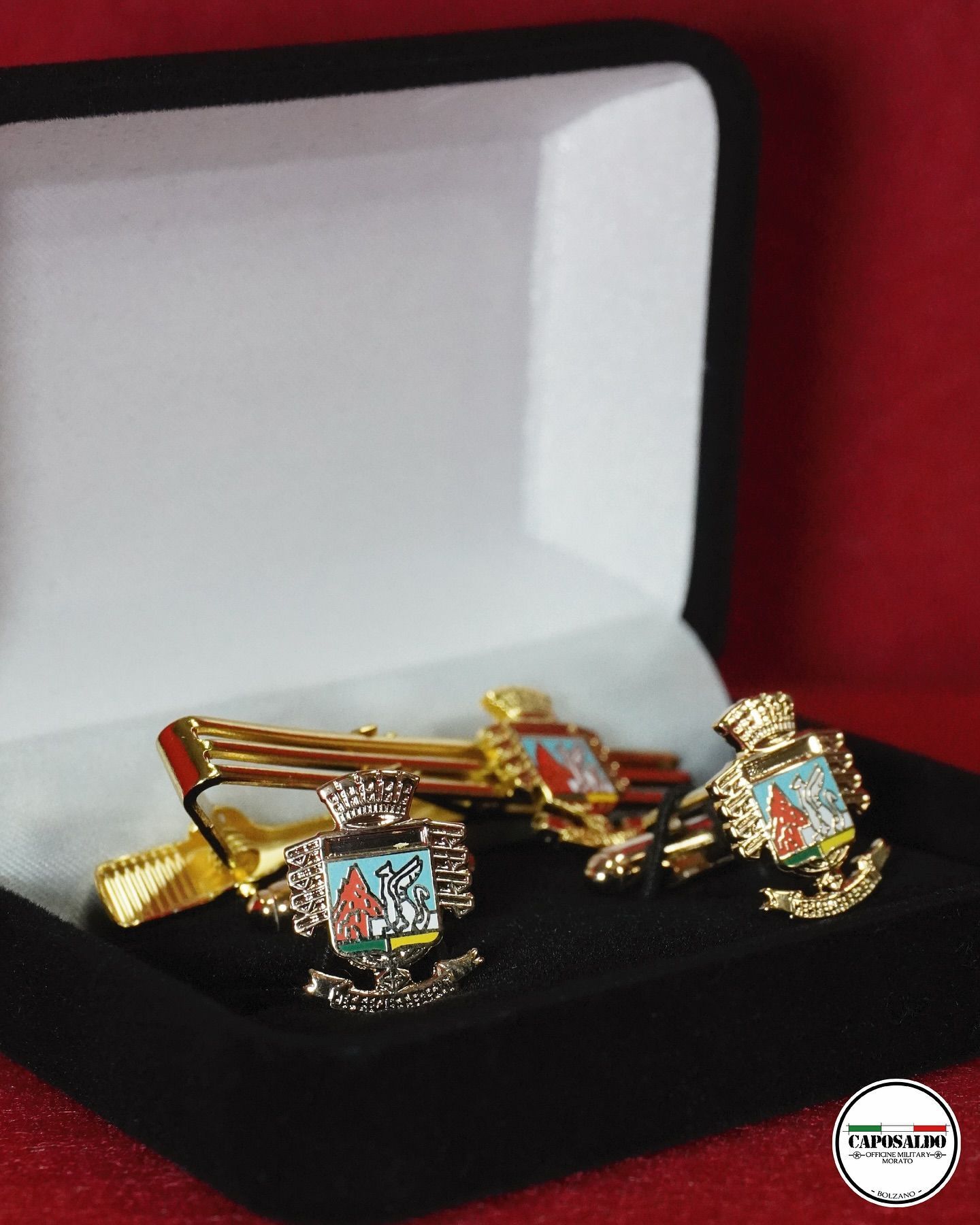 Braccialetti d'oro con ciondoli quadrati colorati esposti in un portagioie nero su sfondo rosso.