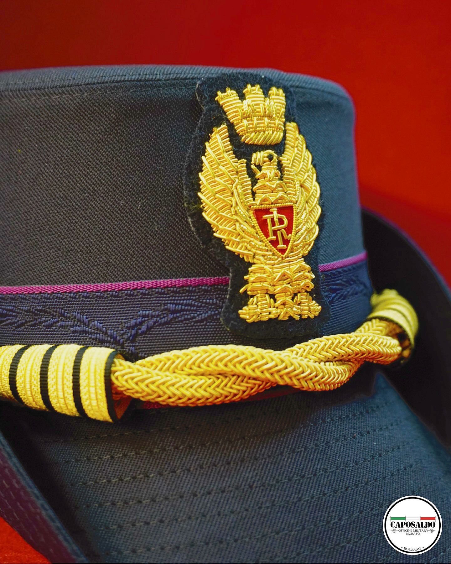Primo piano di un berretto militare nero con treccia dorata e stemma dorato ricamato su sfondo rosso.