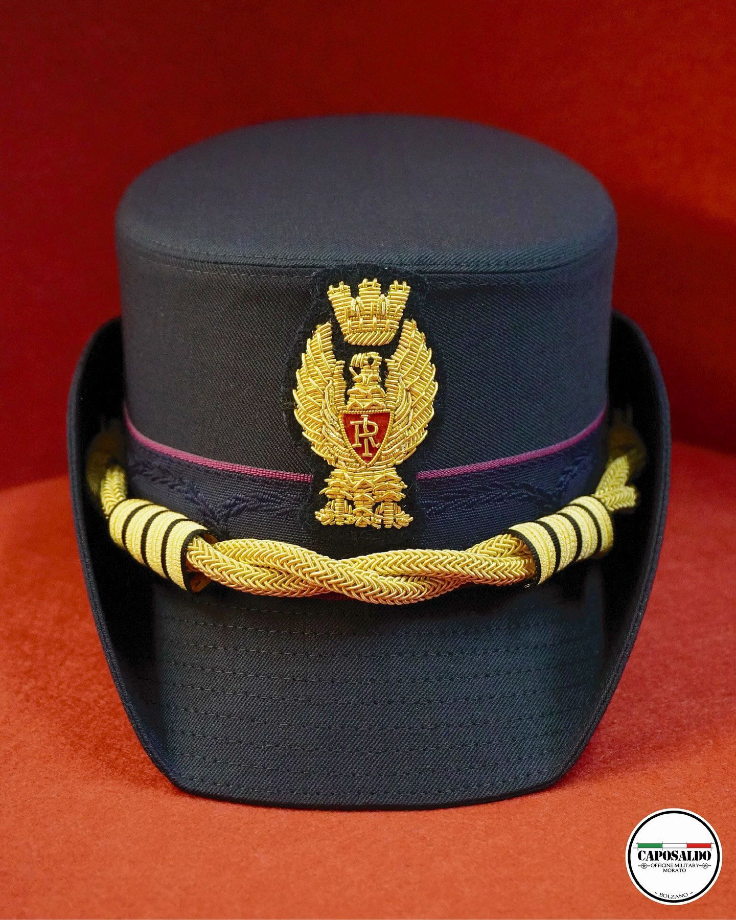 Berretto militare blu scuro con emblema dorato e cordino intrecciato su sfondo rosso.