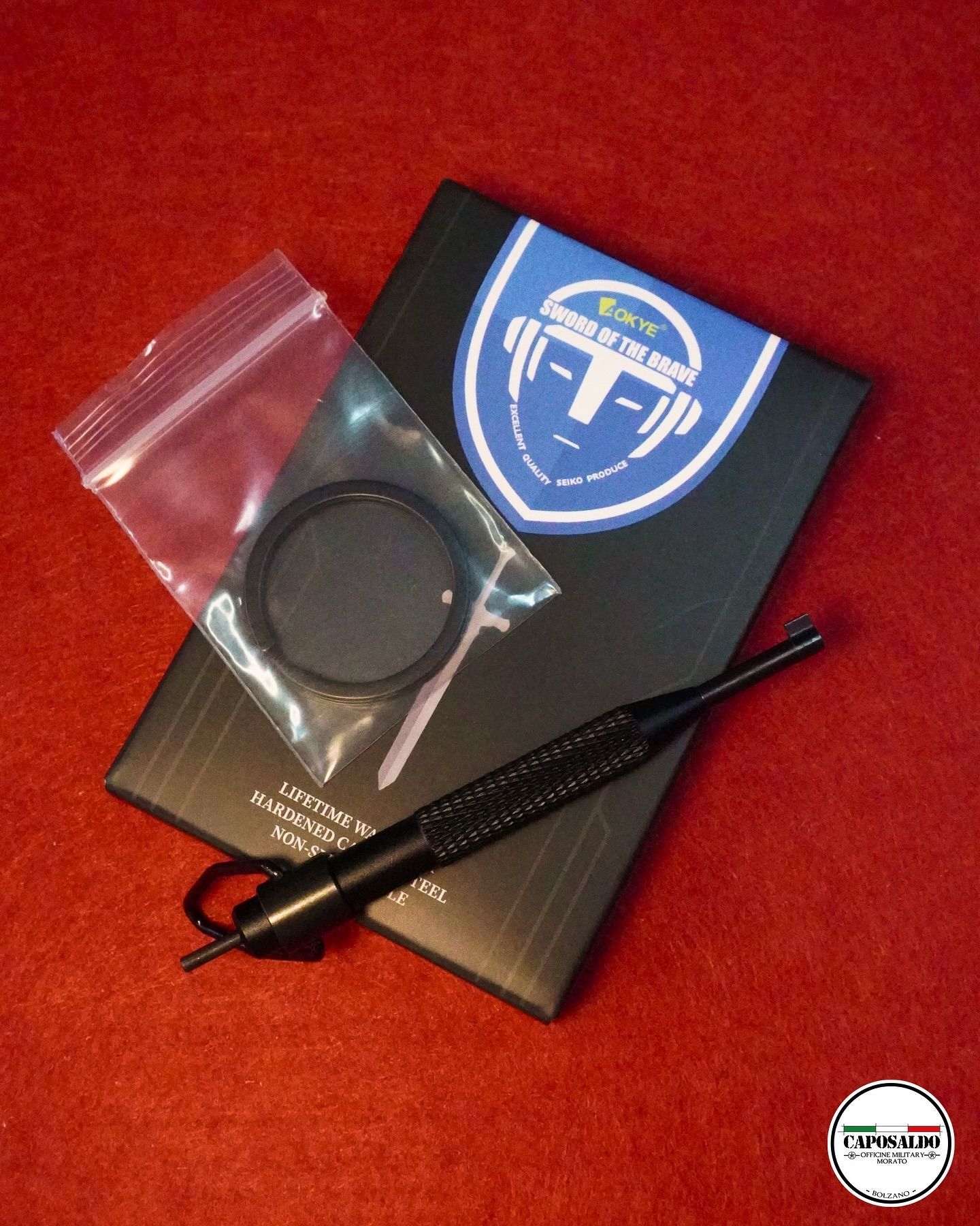 Dispositivo nero con logo blu, antenna e piccolo anello in bustina con zip su sfondo di tessuto rosso.