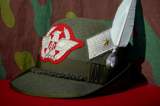 Berretto militare verde oliva con emblema rosso e piuma bianca su sfondo rosso.