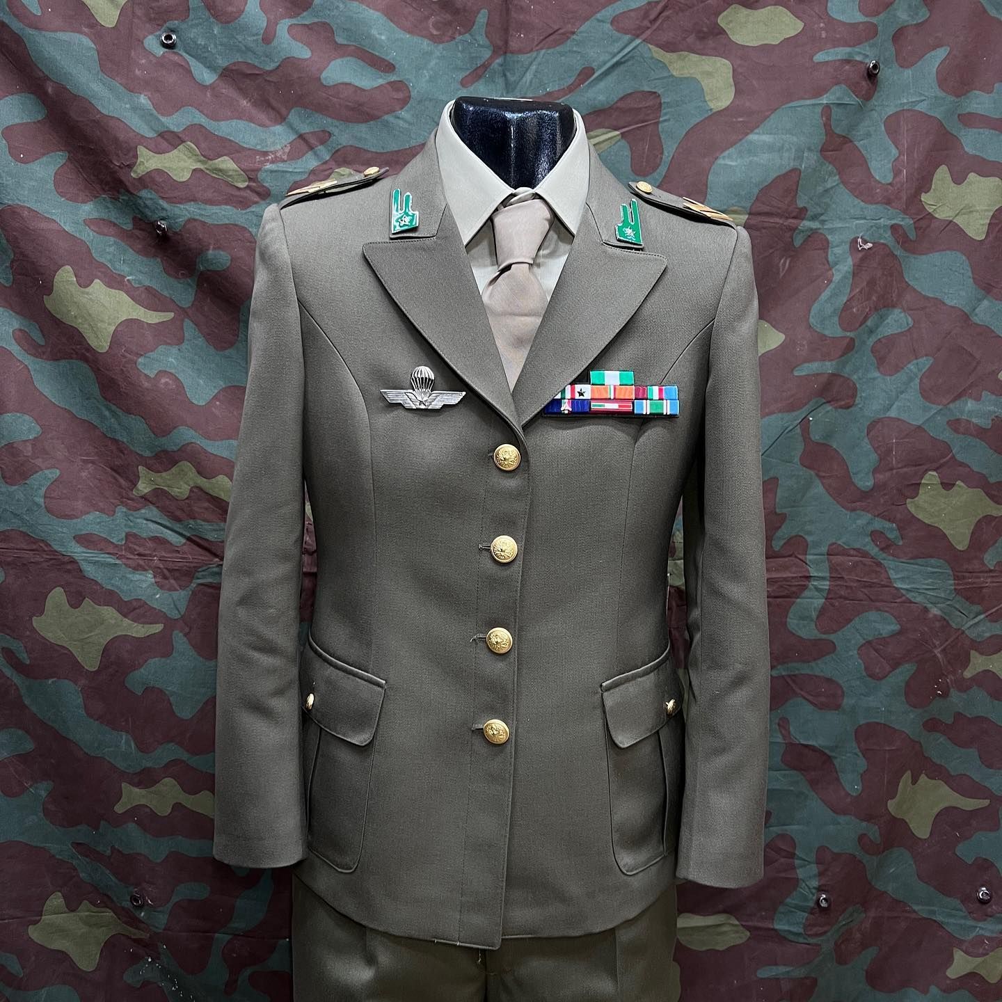 Giacca grigia da uniforme militare con bottoni in ottone e nastrini di servizio su sfondo mimetico.