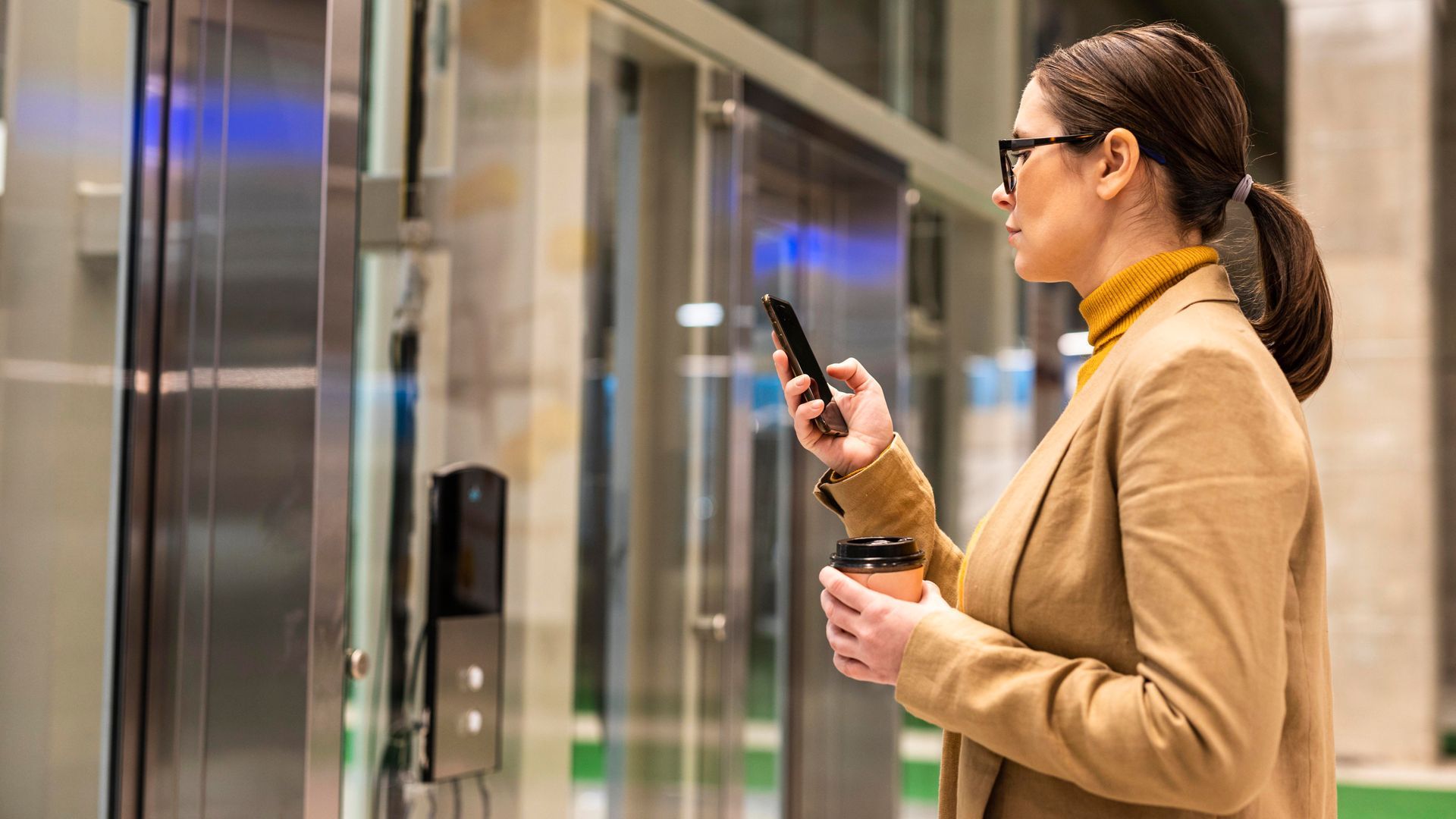 Woman in blazer uses phone near elevator, holding coffee cup.