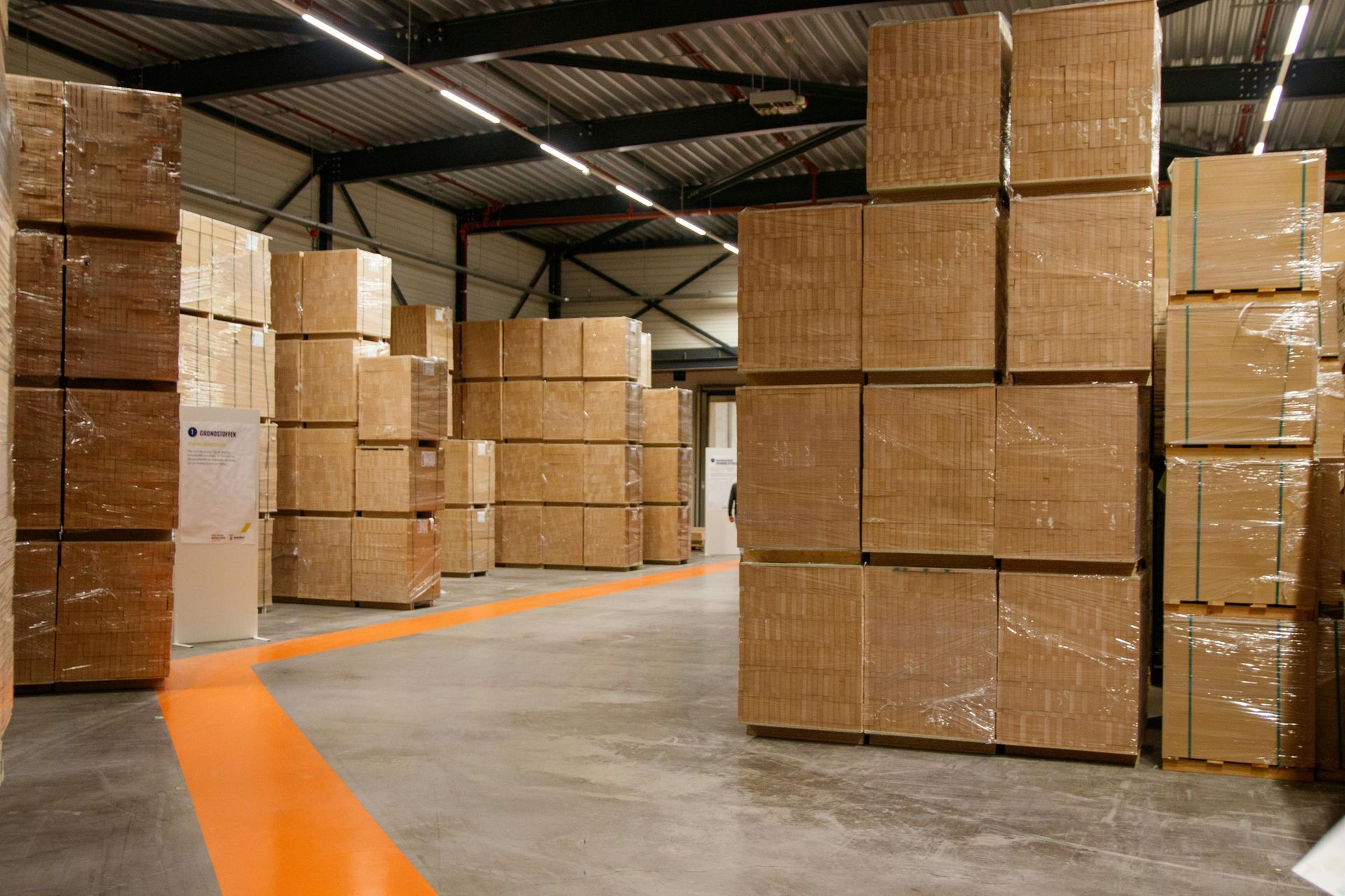 Warehouse interior with stacks of packaged wooden panels and an orange floor marking.