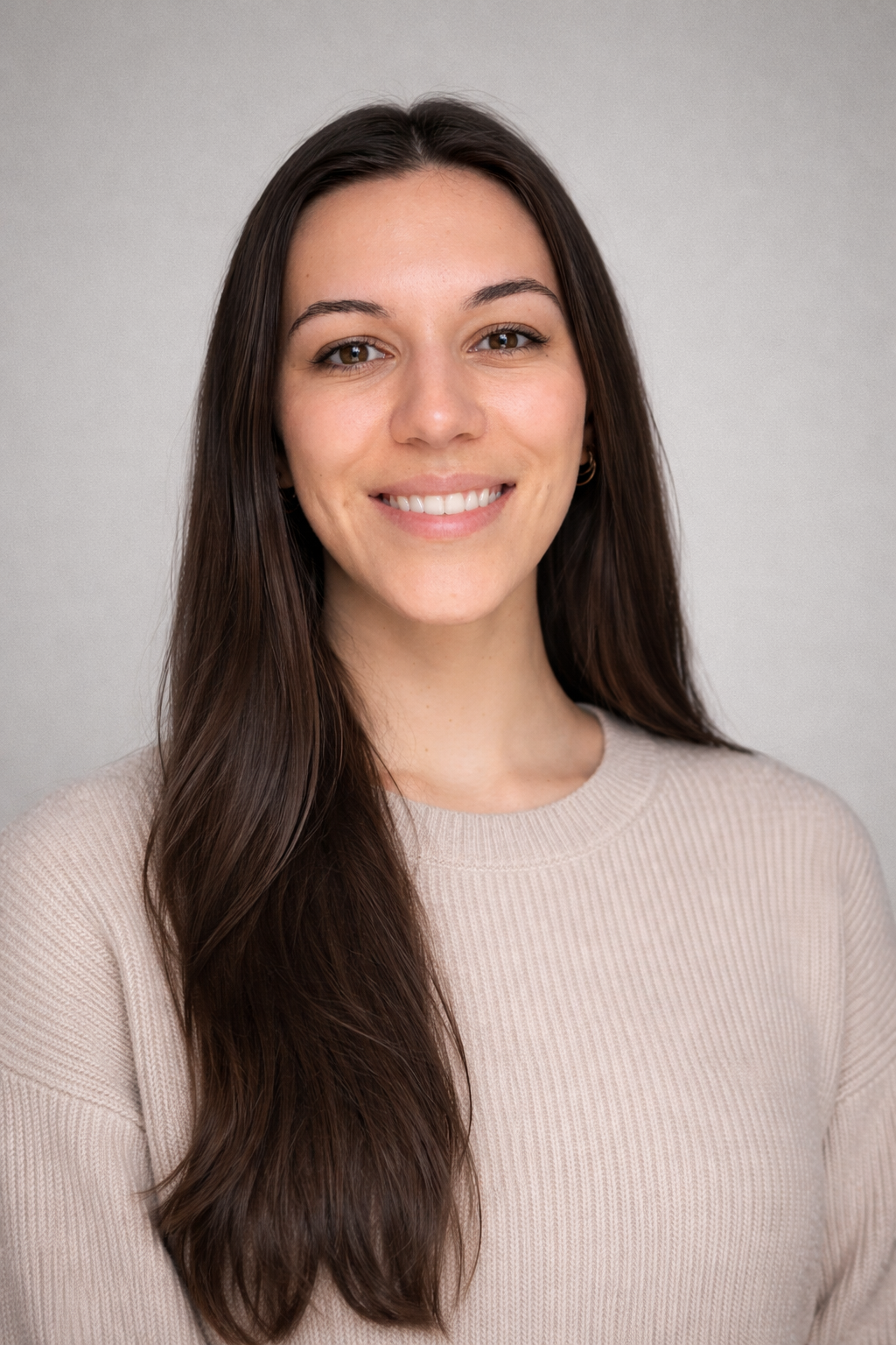Woman with glasses, wearing a purple hat and tan coat, smiles against a gray background.