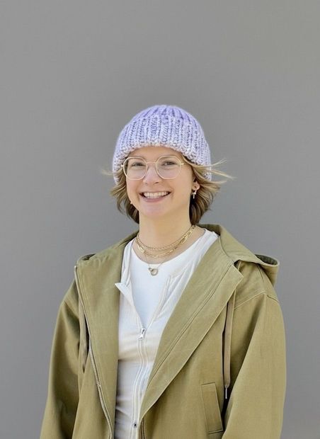 Woman with glasses, wearing a purple hat and tan coat, smiles against a gray background.