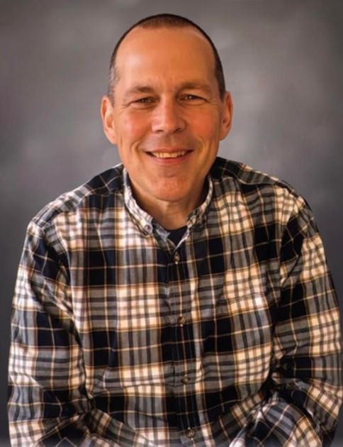 Man with short hair smiling, wearing a plaid shirt, against a gray backdrop.