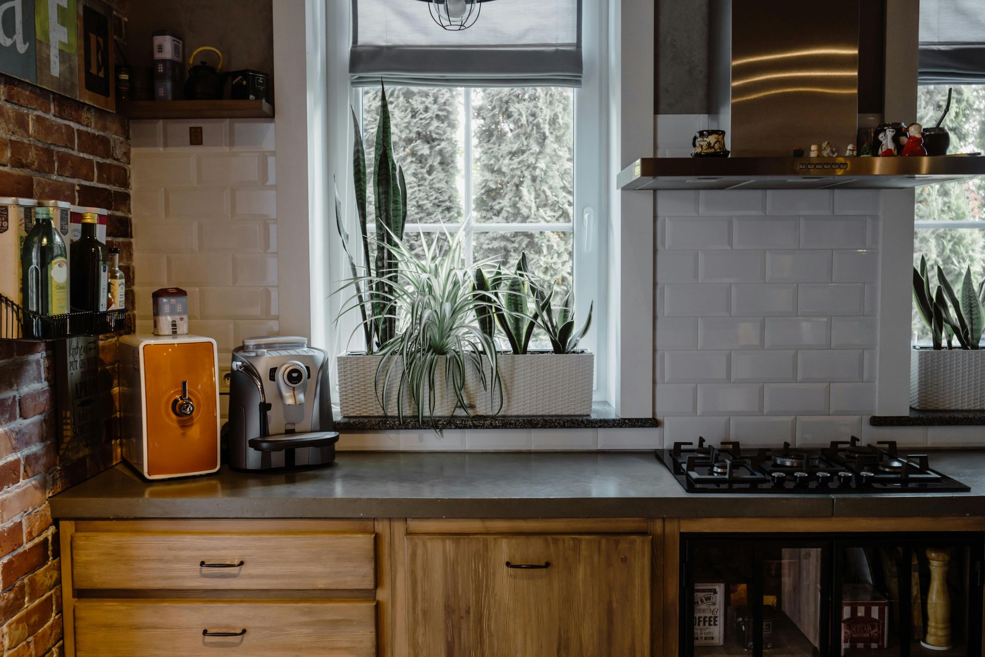 Kitchen with brick wall, light wood cabinets, counter with appliances, plants in window.