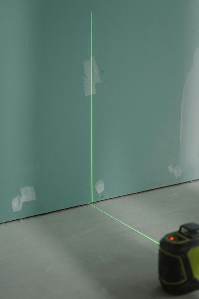 Green laser level projecting a vertical line on a teal wall. The device sits on a concrete floor.