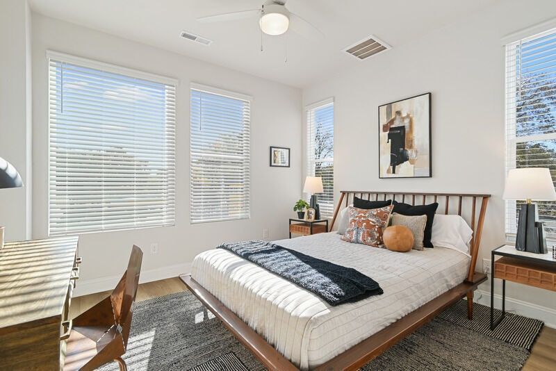 Bedroom with bed, desk, artwork, and multiple windows with blinds.