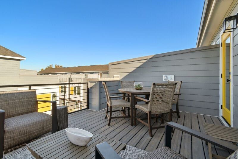 Outdoor deck with seating: table, chairs, and cushioned armchairs. Wooden deck, gray siding, and blue sky.