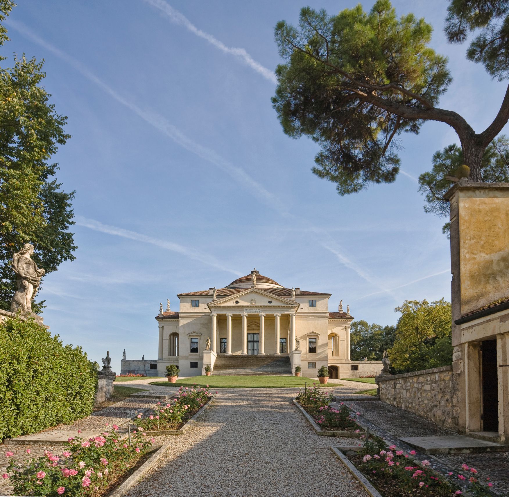Grande villa neoclassica con colonne in fondo a un vialetto di ghiaia, incorniciata da alberi e cielo azzurro.