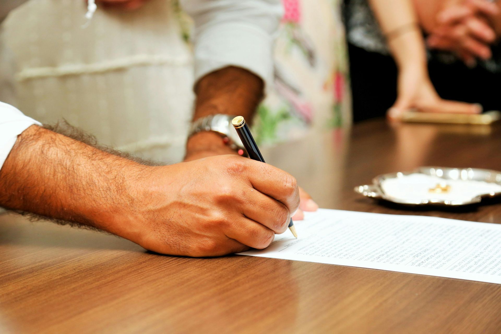 Una mano che firma un documento su un tavolo di legno, con una penna e un piatto sullo sfondo.