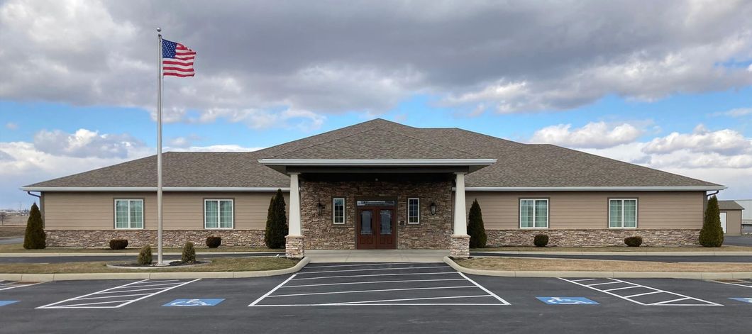 A one-story building with American flag, a blank sign, and a long driveway on a sunny day.