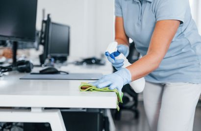 A woman is cleaning a desk with a spray bottle and a cloth.