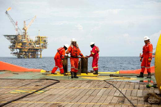 Oil rig workers in orange coveralls on an offshore platform, with a drilling rig in the background.