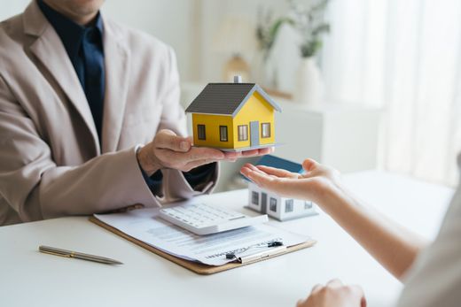 Person in a beige blazer hands a small yellow house model to another person over a desk with paperwork.