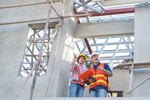 Two construction workers in safety vests and hard hats standing on scaffolding at a building site