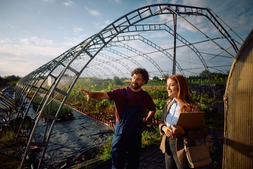 Two people standing by a large arched metal greenhouse frame outdoors, one pointing toward the landscape