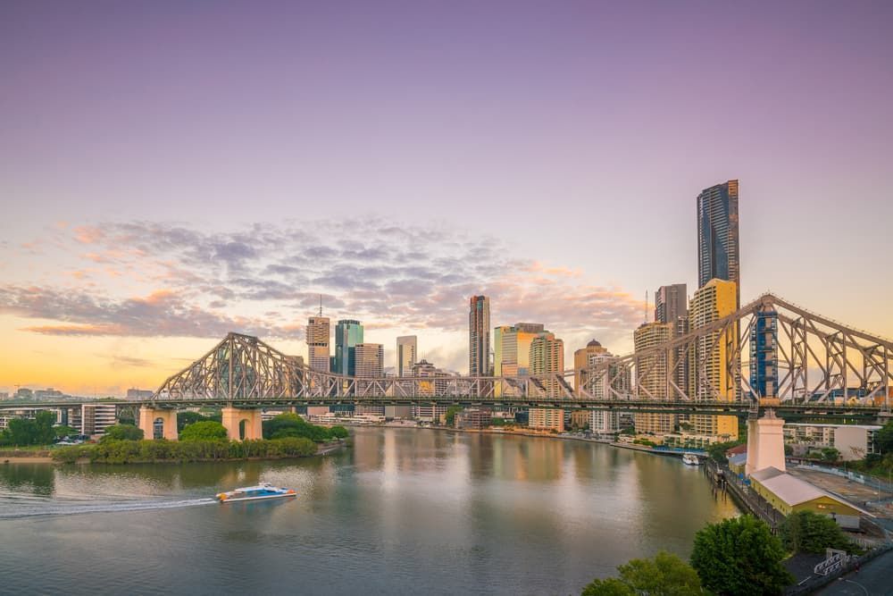 A Boat Is Going Under a Bridge Over a River with A City Skyline in The Background — Lite Moves Furniture Removals In Bonville, NSW