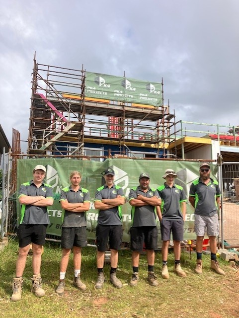 Construction workers in grey shirts and shorts stand in front of scaffolding and a building site — PK4 Projects in Port Macquarie, NSW