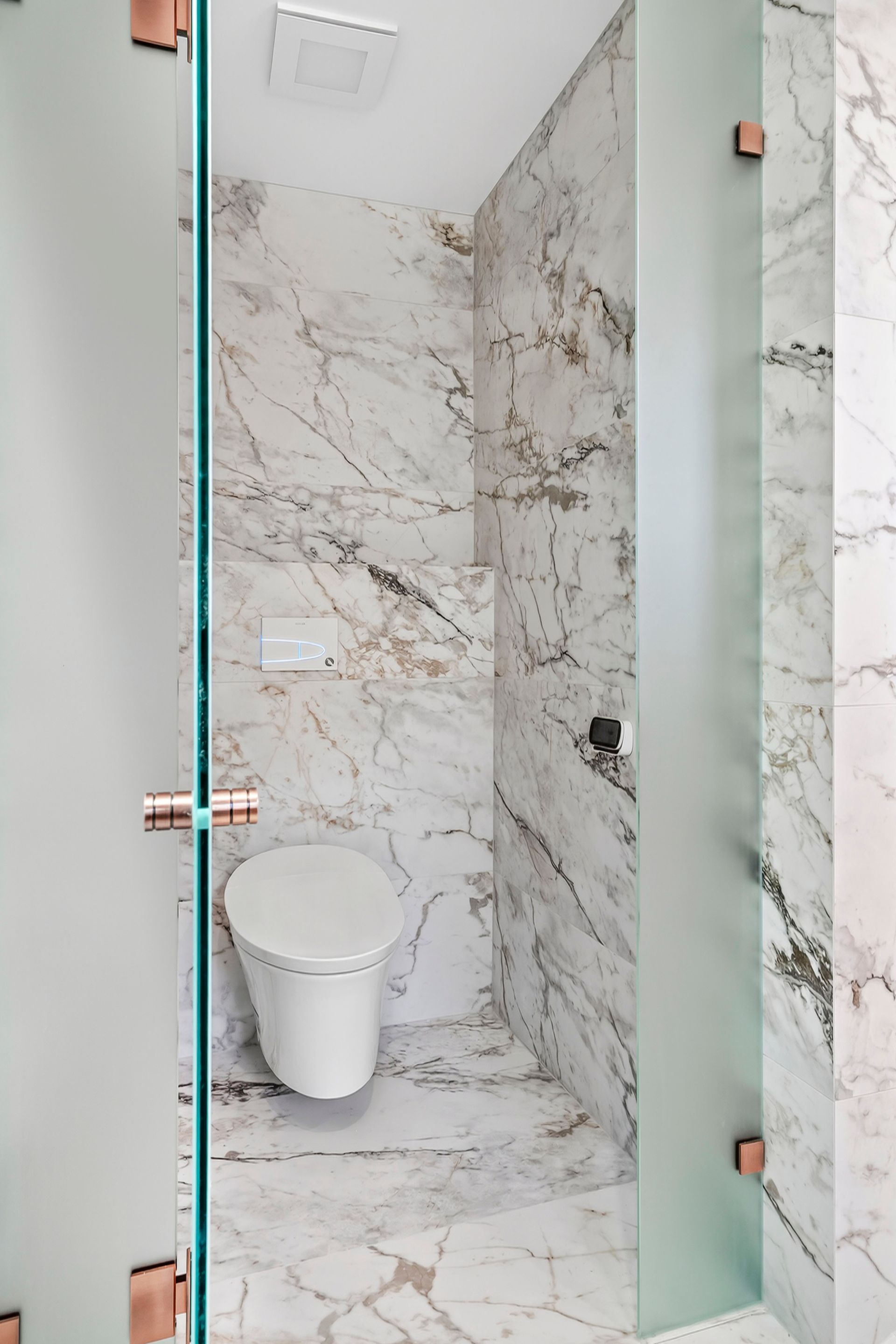 Small, marble-tiled bathroom with a toilet. Frosted glass doors with copper-colored hardware — PK4 Projects in Port Macquarie, NSW