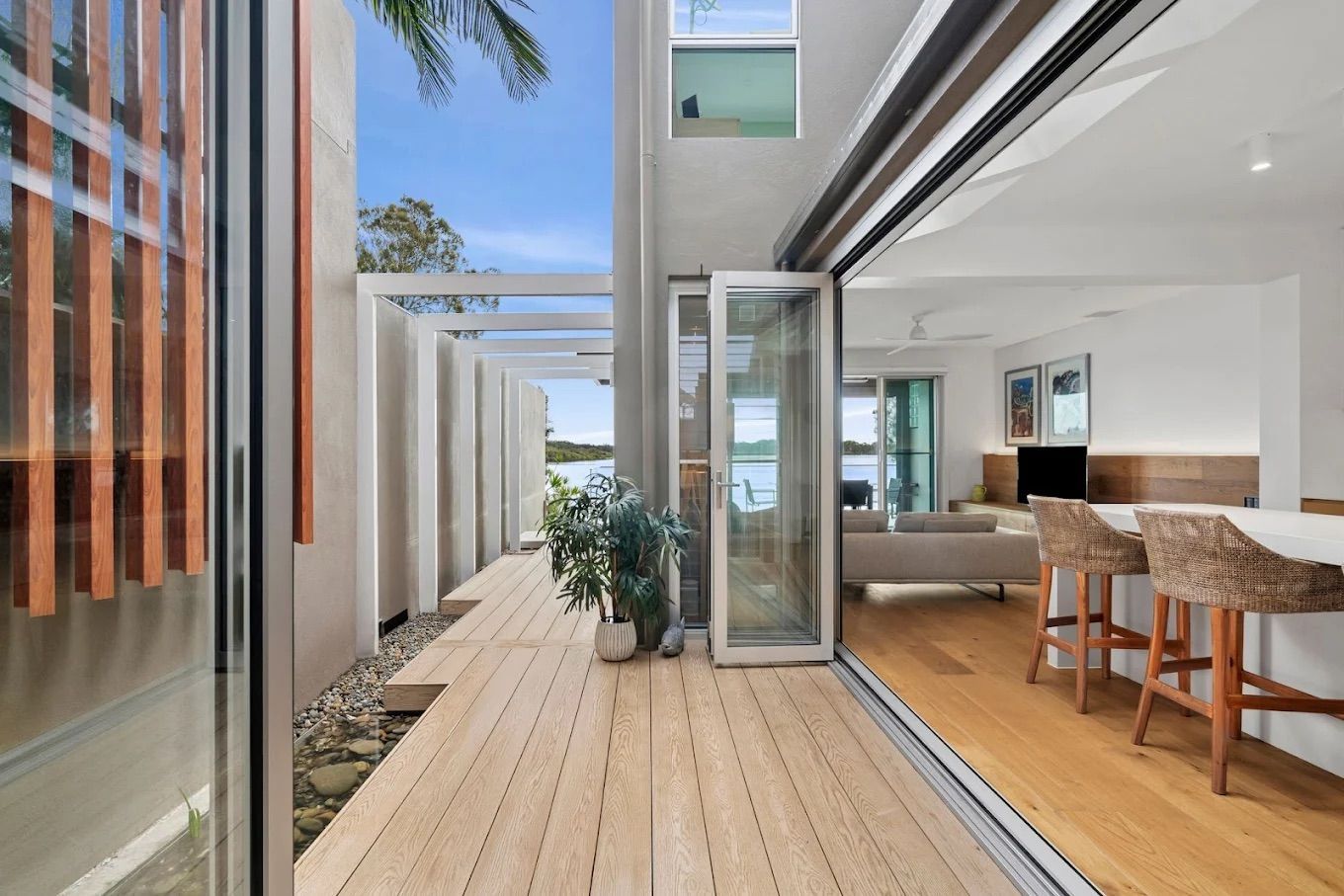 Deck with wooden floor extends from open glass doors to an outdoor area with a view of water and a white-walled structure — PK4 Projects in Port Macquarie, NSW 