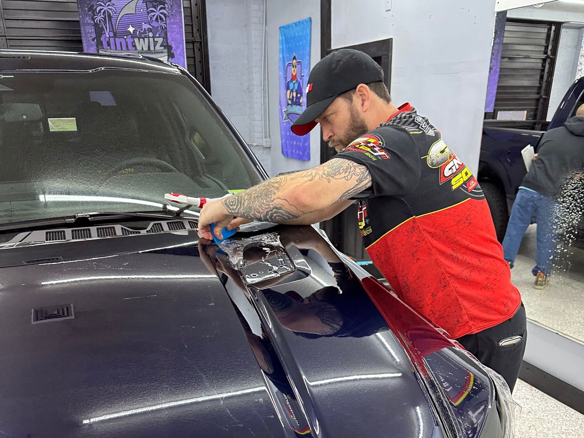 Man in a black and red shirt applying film to a car's hood. Indoors, well lit.