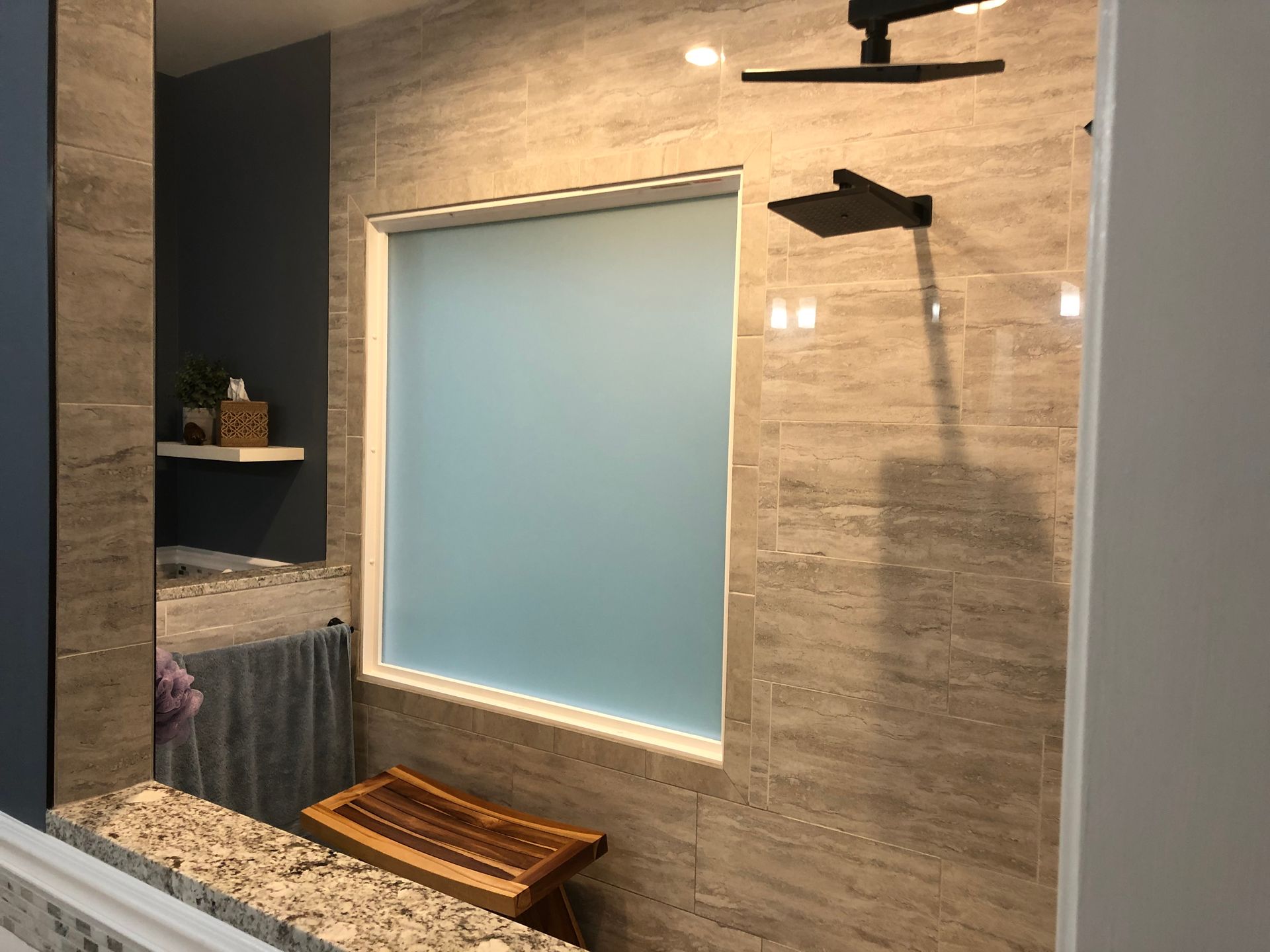 Shower with stone-like tile, frosted window, showerhead, wooden bench, and a small shelf.