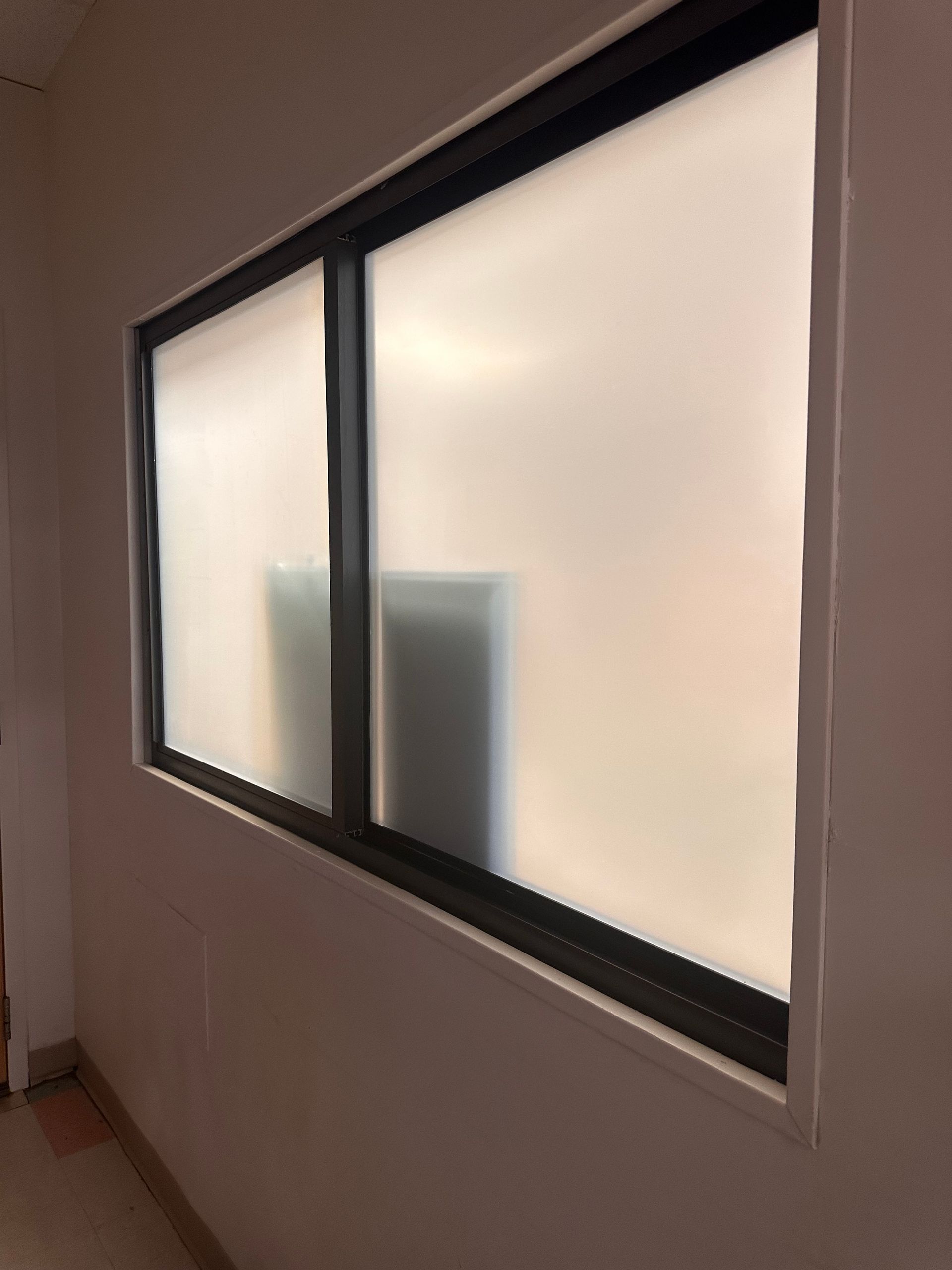 Frosted-glass window in a white-walled room, with a black frame. The interior of the room is dimly visible.