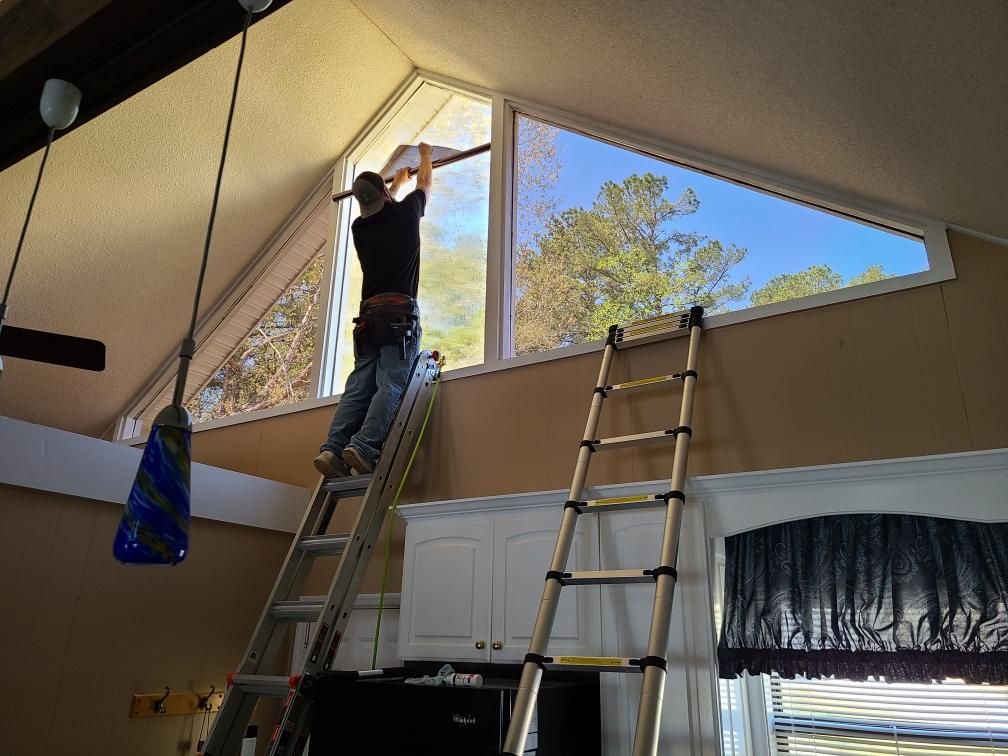 Person installing window film on a triangular window, standing on a ladder inside a house.