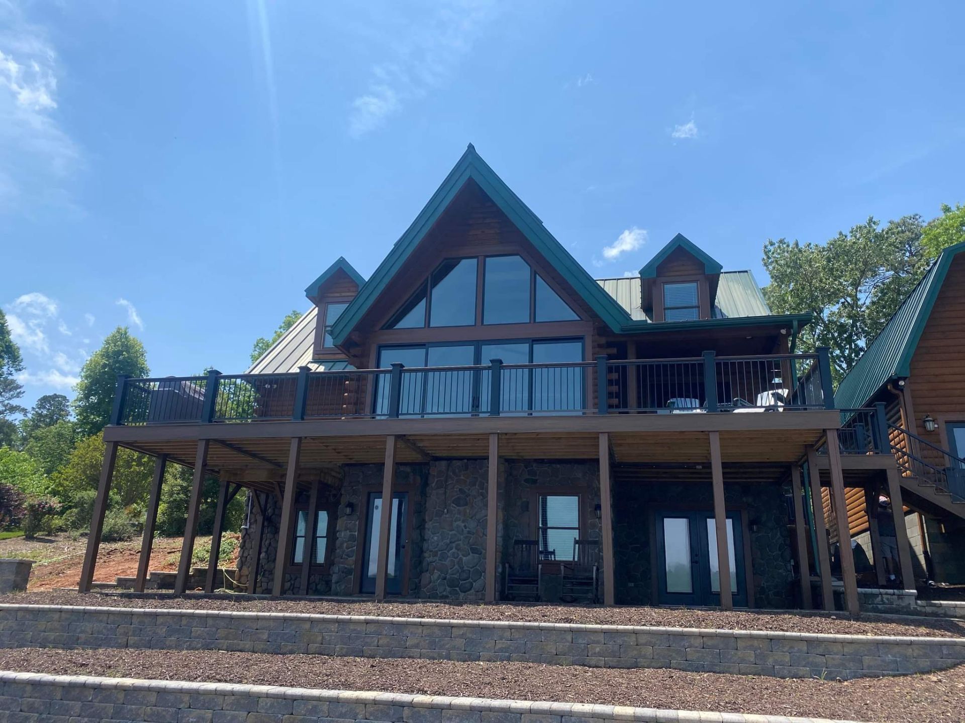Two-story log cabin with a large deck and tall windows, built on a stone foundation, under a blue sky.