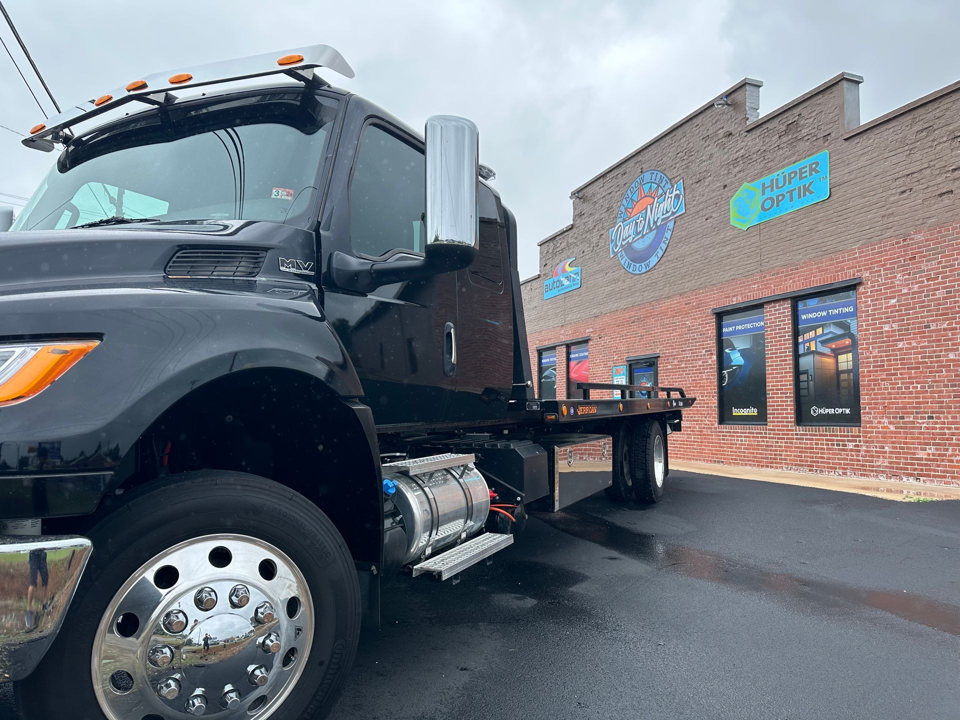 Black tow truck parked in front of a brick building with a logo on the wall. Cloudy day, wet pavement.