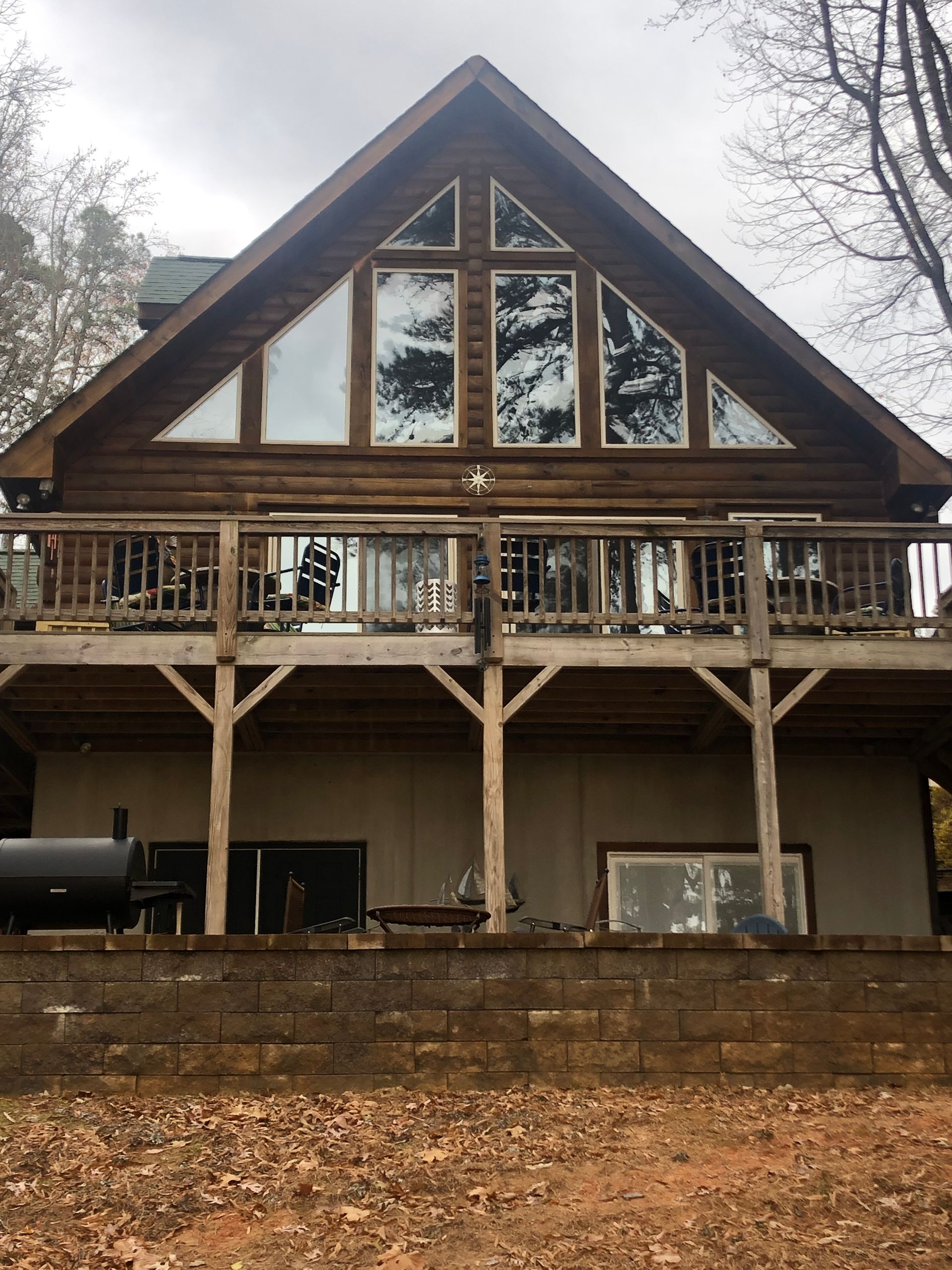 A wooden cabin with a large deck and triangular windows. A grill is on the ground level. The sky is overcast.